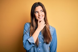 Young beautiful girl wearing casual denim shirt standing over isolated yellow background with hand on chin thinking about question, pensive expression. Smiling with thoughtful face. Doubt concept.