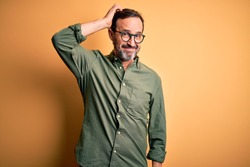 Middle age hoary man wearing casual green shirt and glasses over isolated yellow background confuse and wonder about question. Uncertain with doubt, thinking with hand on head. Pensive concept.
