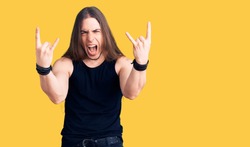 Young adult man with long hair wearing goth style with black clothes shouting with crazy expression doing rock symbol with hands up. music star. heavy music concept. 