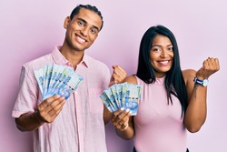 Young latin couple holding south african rand banknotes screaming proud, celebrating victory and success very excited with raised arm 