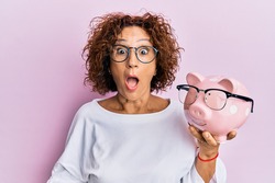 Beautiful middle age mature woman holding piggy bank with glasses scared and amazed with open mouth for surprise, disbelief face 