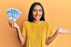 Young brunette woman holding south african 100 rand banknotes celebrating achievement with happy smile and winner expression with raised hand 