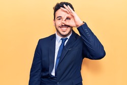 Young hispanic man wearing suit doing ok gesture with hand smiling, eye looking through fingers with happy face. 