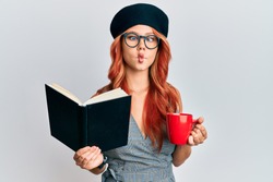 Young redhead woman wearing french beret reading a book and drinking a coffee making fish face with mouth and squinting eyes, crazy and comical. 