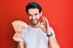 Young hispanic man holding mexican pesos doing ok sign with fingers, smiling friendly gesturing excellent symbol 