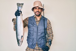 Handsome hispanic man with beard holding fishing rod and raw salmon smiling with a happy and cool smile on face. showing teeth. 