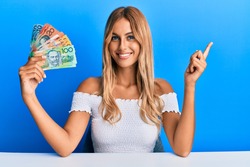 Beautiful blonde young woman holding australian dollars smiling happy pointing with hand and finger to the side 