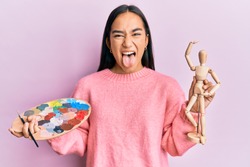 Young asian woman holding painter palette and art manikin sticking tongue out happy with funny expression. 