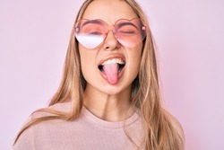 Young beautiful blonde woman wearing heart shaped sunglasses sticking tongue out happy with funny expression. 