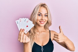 Beautiful caucasian blonde girl holding ace poker cards smiling happy and positive, thumb up doing excellent and approval sign 