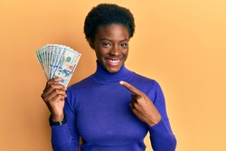 Young african american girl holding dollars smiling happy pointing with hand and finger 