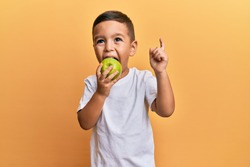 Adorable latin toddler smiling happy eating green apple looking to the camera over isolated yellow background.