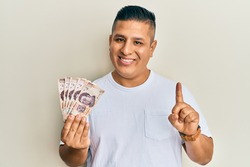 Young latin man holding 500 mexican pesos banknotes smiling with an idea or question pointing finger with happy face, number one 