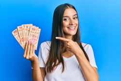 Young hispanic woman holding mexican pesos smiling happy pointing with hand and finger 