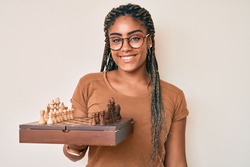 Young african american woman with braids holding chess board wearing glasses smiling with a happy and cool smile on face. showing teeth. 