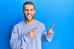 Young caucasian man pointing with fingers to the side sticking tongue out happy with funny expression. 