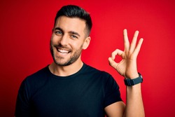 Young handsome man wearing casual black t-shirt standing over isolated red background smiling positive doing ok sign with hand and fingers. Successful expression.