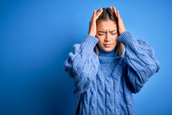 Young beautiful blonde woman wearing winter wool sweater over blue isolated background suffering from headache desperate and stressed because pain and migraine. Hands on head.