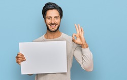 Young hispanic man holding blank empty banner doing ok sign with fingers, smiling friendly gesturing excellent symbol 
