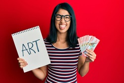 Beautiful asian young woman holding art notebook and 100 new zealand dollars sticking tongue out happy with funny expression. 