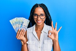 Beautiful hispanic woman holding south african 100 rand banknotes doing ok sign with fingers, smiling friendly gesturing excellent symbol 