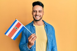 Young arab man holding costa rica flag looking positive and happy standing and smiling with a confident smile showing teeth 