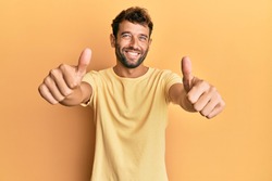 Handsome man with beard wearing casual yellow tshirt over yellow background approving doing positive gesture with hand, thumbs up smiling and happy for success. winner gesture. 