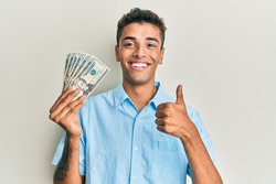 Young handsome african american man holding 20 dollars banknotes smiling happy and positive, thumb up doing excellent and approval sign 