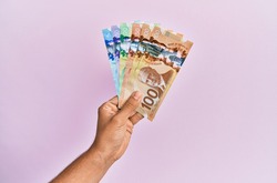 Hispanic hand holding canadian dollars banknotes over isolated pink background.