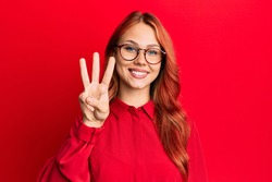 Young beautiful redhead woman wearing casual clothes and glasses over red background showing and pointing up with fingers number three while smiling confident and happy. 