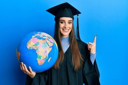 Beautiful brunette young woman wearing graduation robe holding world ball smiling happy pointing with hand and finger to the side 