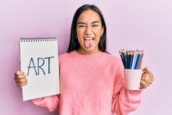 Young asian woman holding art notebook and colored pencils sticking tongue out happy with funny expression. 