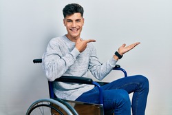 Young hispanic man sitting on wheelchair amazed and smiling to the camera while presenting with hand and pointing with finger. 