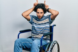 Handsome hispanic man sitting on wheelchair doing funny gesture with finger over head as bull horns 