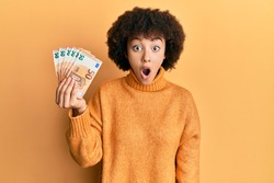Young hispanic girl holding bunch of 50 euro banknotes scared and amazed with open mouth for surprise, disbelief face 