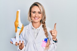 Beautiful young blonde doctor woman holding anatomical model of knee joint smiling with an idea or question pointing finger with happy face, number one 
