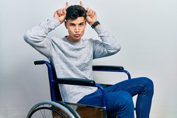 Young hispanic man sitting on wheelchair doing funny gesture with finger over head as bull horns 