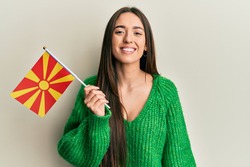 Young hispanic girl holding macedonian flag looking positive and happy standing and smiling with a confident smile showing teeth 