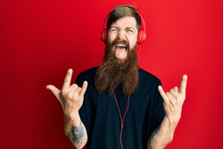 Redhead man with long beard listening to music using headphones shouting with crazy expression doing rock symbol with hands up. music star. heavy concept. 