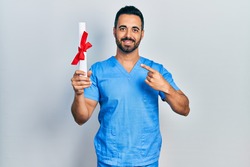 Handsome hispanic man with beard wearing blue male nurse uniform holding diploma smiling happy pointing with hand and finger 