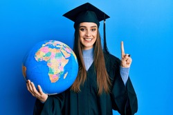 Beautiful brunette young woman wearing graduation robe holding world ball smiling with an idea or question pointing finger with happy face, number one 
