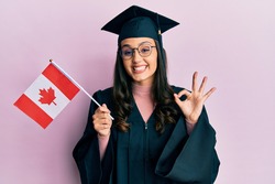 Young hispanic woman wearing graduation uniform holding canada flag doing ok sign with fingers, smiling friendly gesturing excellent symbol 