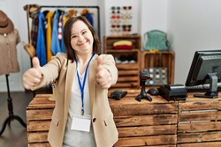 Young down syndrome woman working as manager at retail boutique approving doing positive gesture with hand, thumbs up smiling and happy for success. winner gesture. 