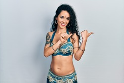 Young woman wearing bindi and traditional belly dance clothes pointing to the back behind with hand and thumbs up, smiling confident 