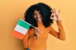 African american woman with afro hair holding ireland flag doing ok sign with fingers, smiling friendly gesturing excellent symbol 
