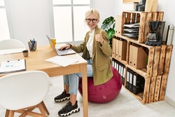 Beautiful caucasian business woman working at the office sitting on pilates ball smiling happy and positive, thumb up doing excellent and approval sign 