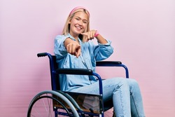 Beautiful blonde woman sitting on wheelchair pointing to you and the camera with fingers, smiling positive and cheerful 