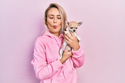 Beautiful young blonde woman hugging cute chihuahua dog making fish face with mouth and squinting eyes, crazy and comical. 