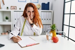 Young redhead woman nutritionist doctor at the clinic hand on mouth telling secret rumor, whispering malicious talk conversation 