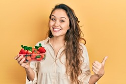 Young hispanic girl holding strawberries pointing to the back behind with hand and thumbs up, smiling confident 